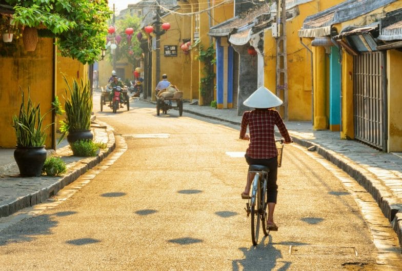 Hoi An (Hoian), Vietnam - April 12, 2018: Vietnamese woman in traditional bamboo hat bicycling along Hoi An Ancient Town. Awesome view of old street decorated with colorful silk lanterns at sunrise.