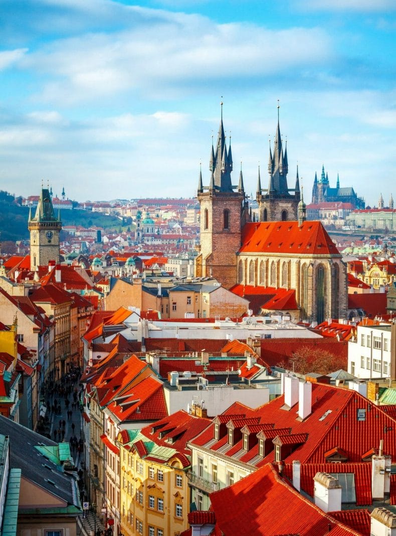 Altas agujas de las torres de la Iglesia de Týn en la ciudad de Praga (Catedral de Nuestra Señora antes de Týn), paisaje urbano panorámico con tejados rojos de casas en la Ciudad Vieja y cielo azul con nubes